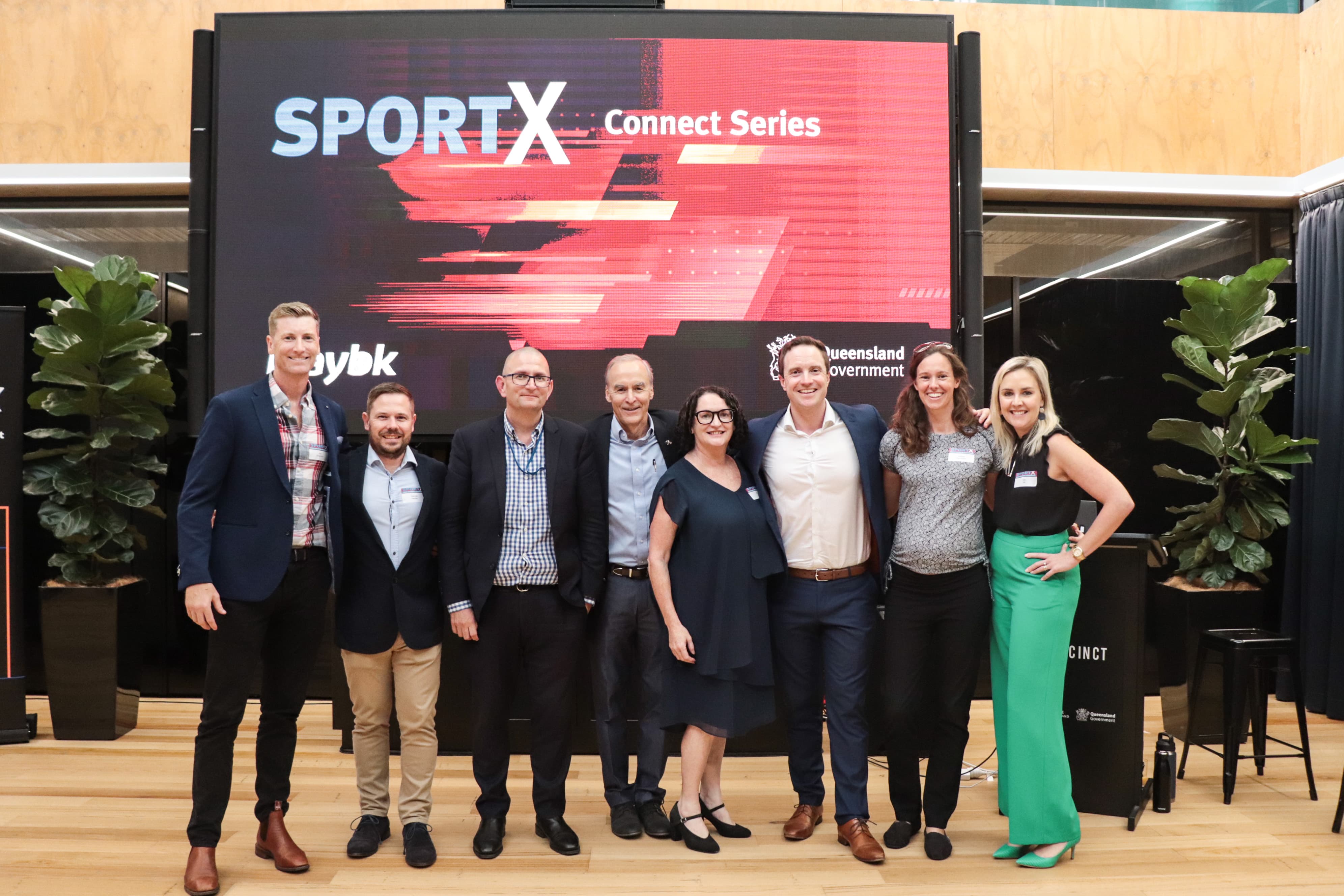 Eight people on a stage in a row smiling at the camera with SportX logo in the background on a large screen