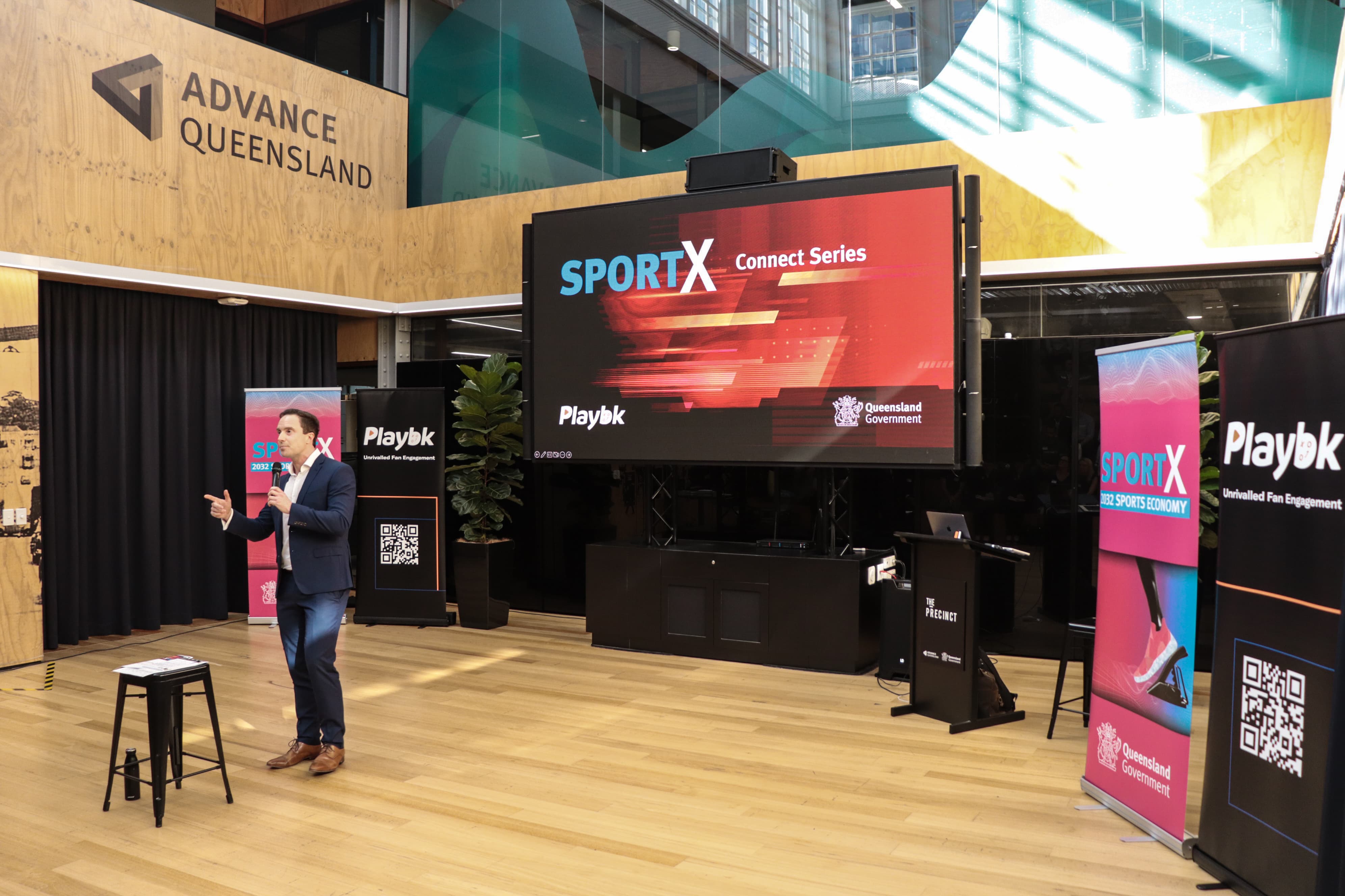Man holding a microphone giving a presentation with a SportX logo in the background on a large screen